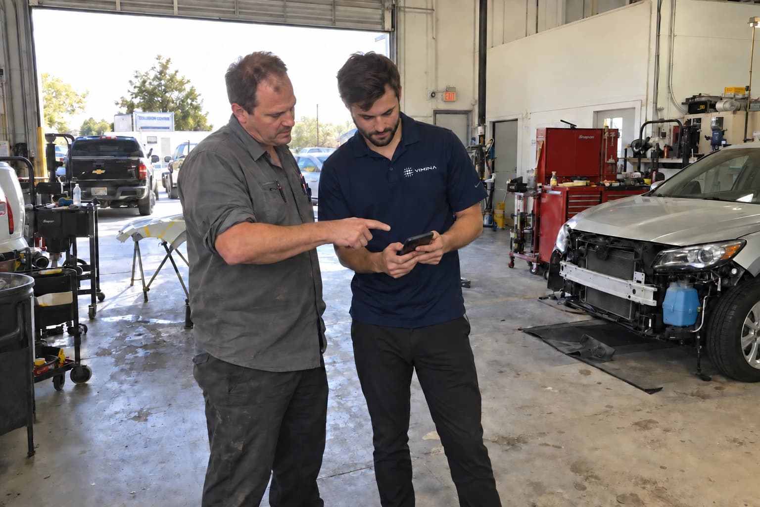 Illustrative scene of a collision shop owner reviewing a phone with a Vimina team member in the service bay